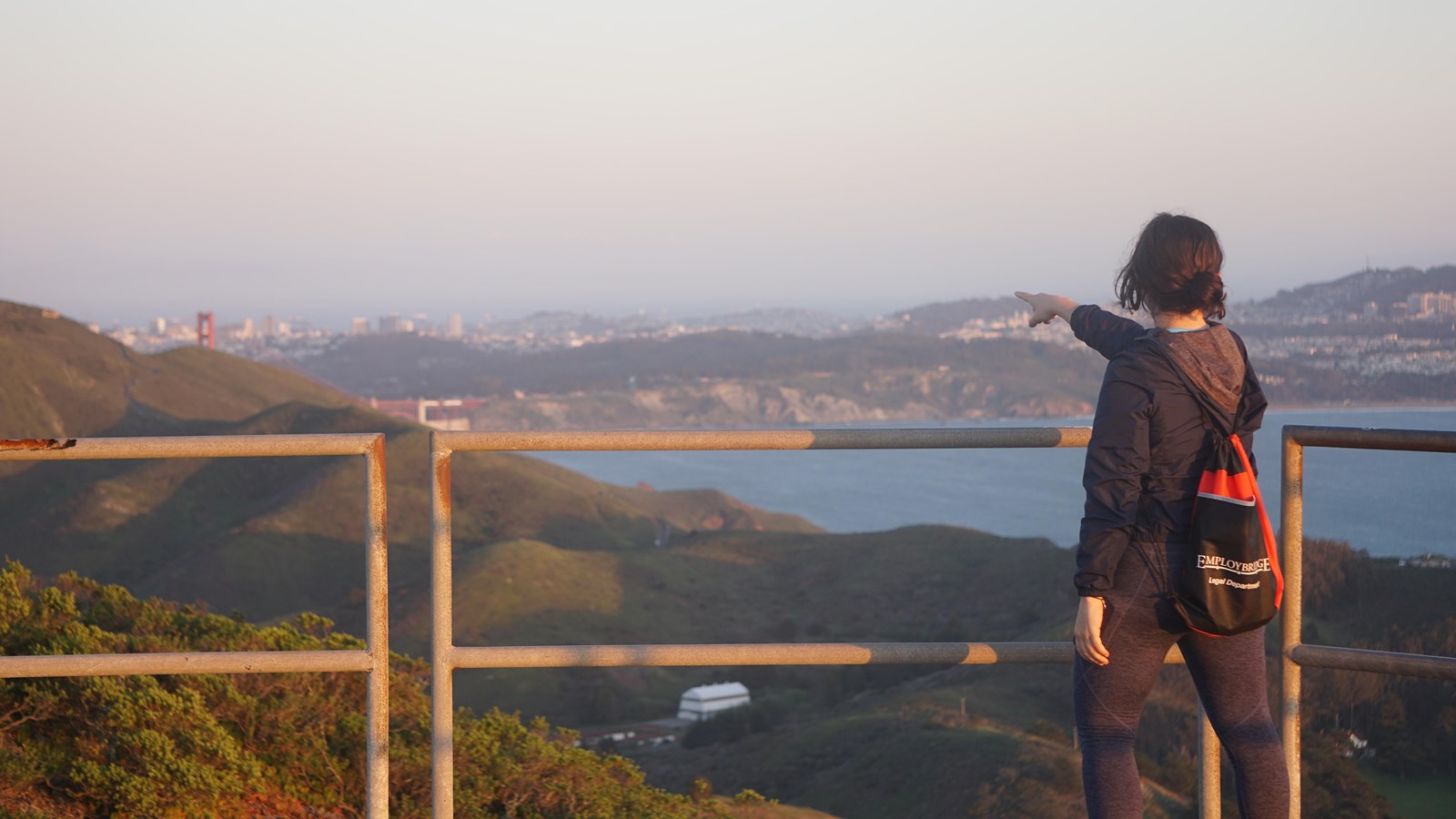 A person stands behind railing at Hill 88 and points to the Golden Gate Bridge in the distance.