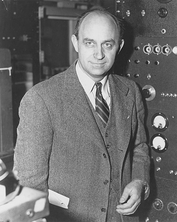 A black and white photo of a man in a suit in front of the camera with scientific equipment behind.