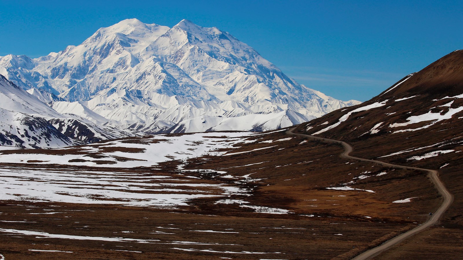 a dirt road leading across a tree-less landscape toward a huge snowy mountain