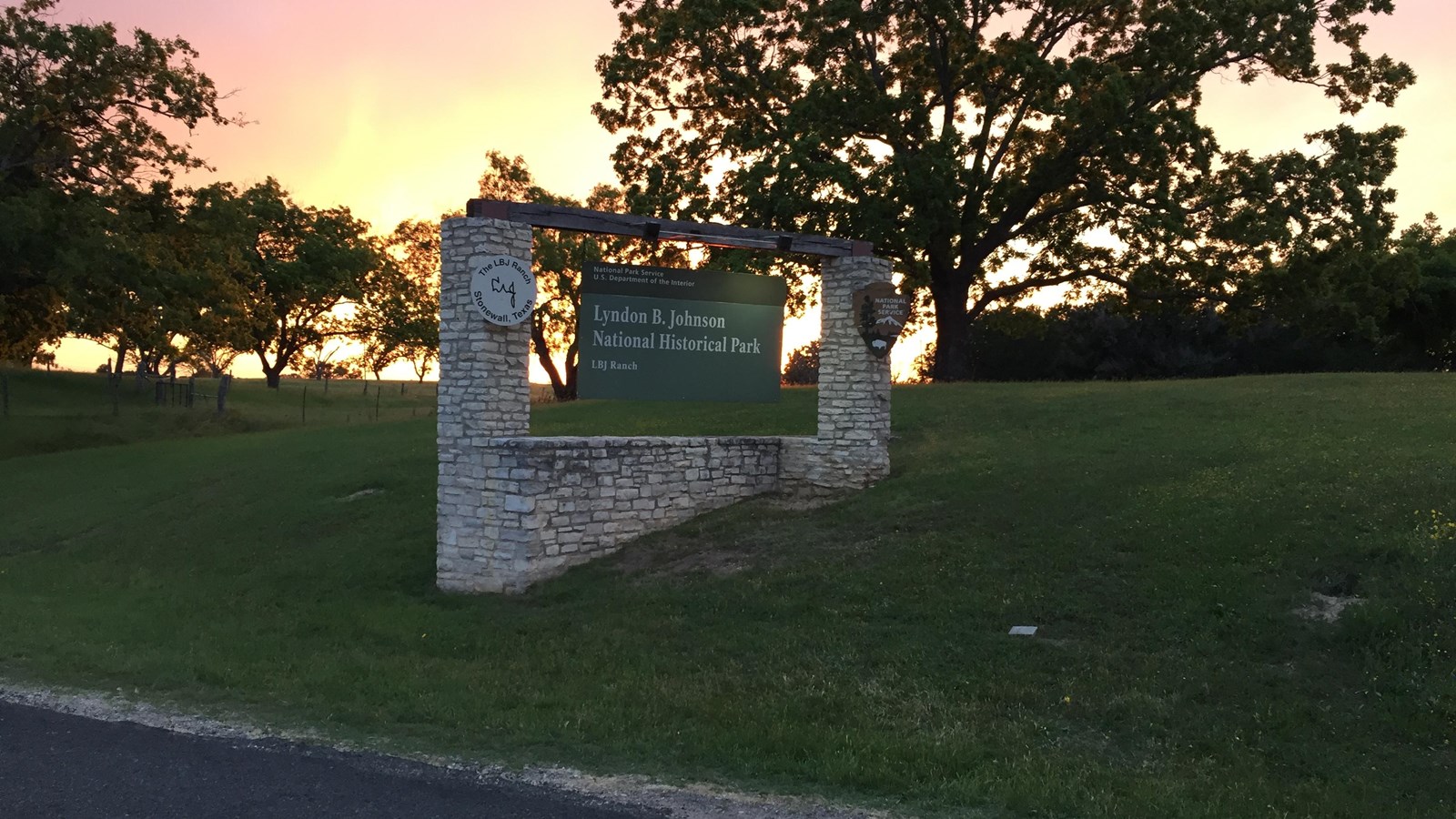 A wood and stone sign is highlighted from behind by a bright yellow, pink, and orange sky.