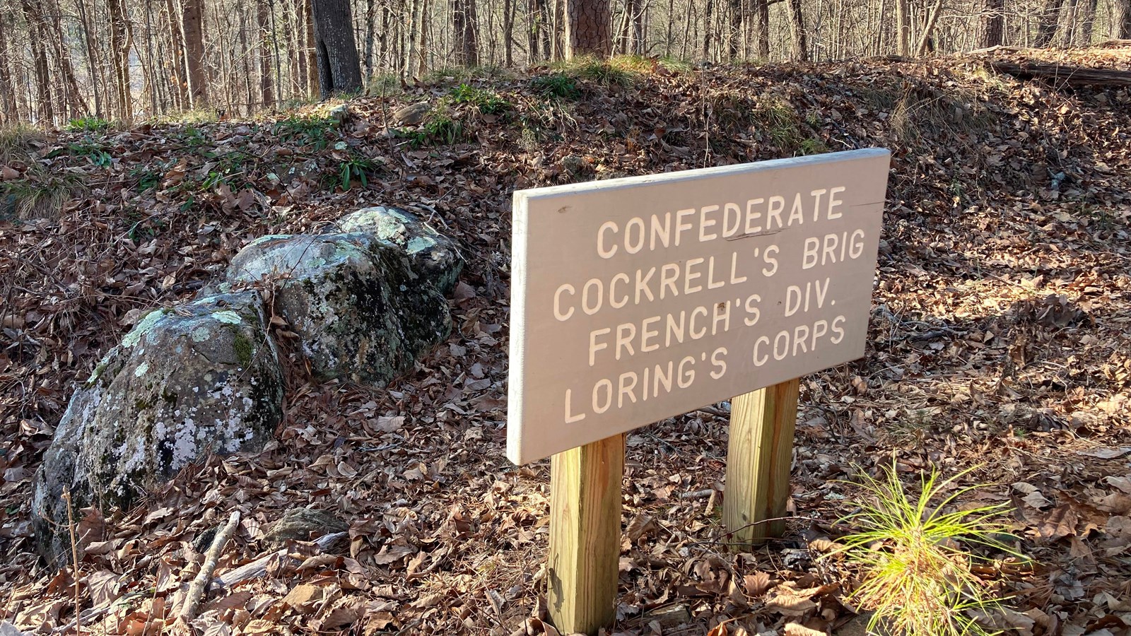 A short gray sign sits in front of a boulder t in the remains of confederate trenches. 