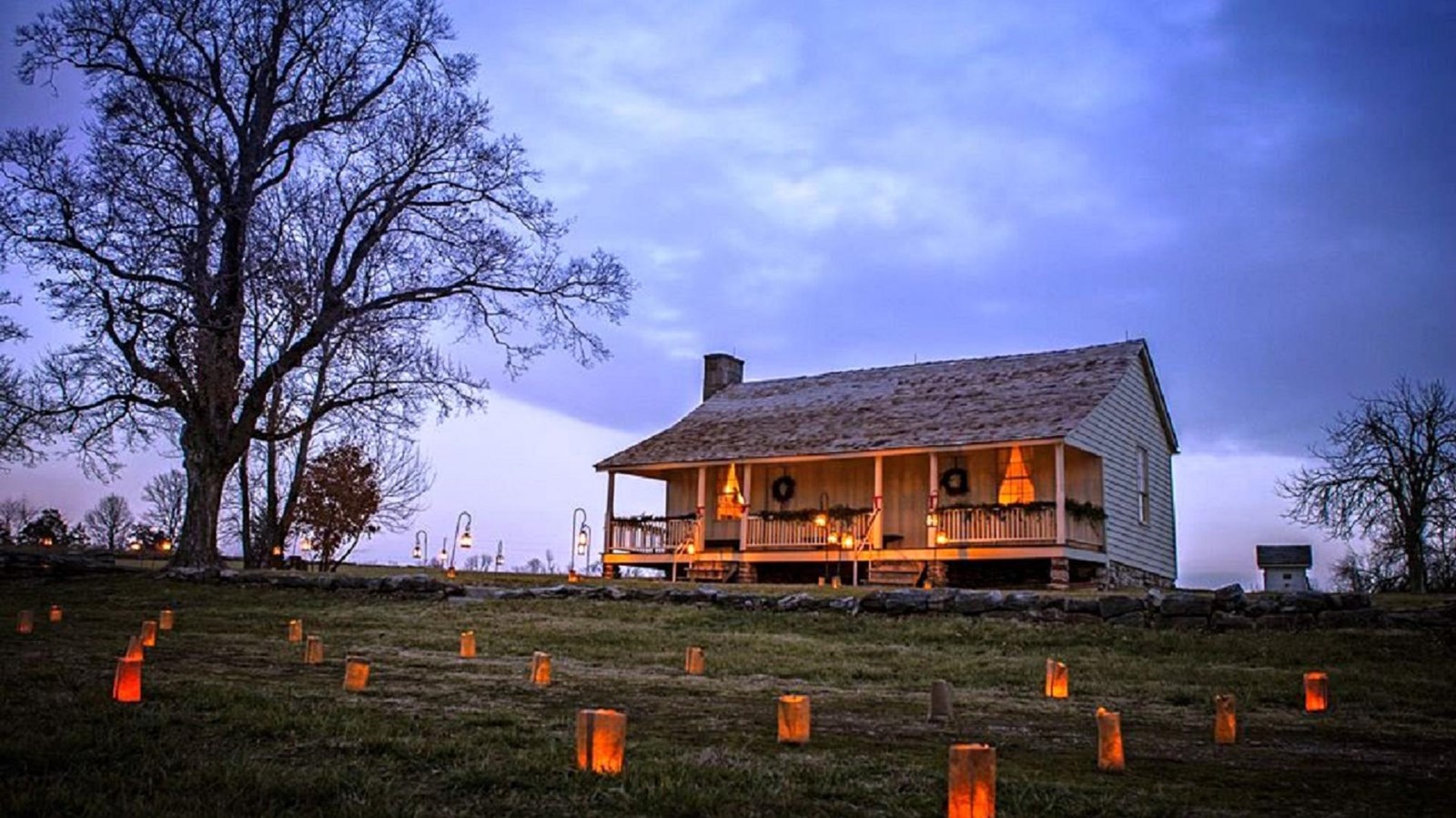 An old house glows in lantern light at dusk with luminaries lit by candles on grassy area out front