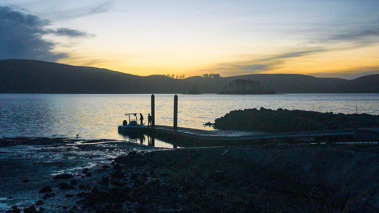 Two persons stand on a floating dock adjacent to their motor boat shortly after sunset.