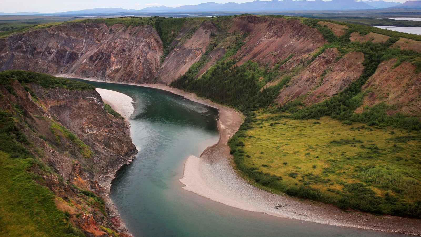 River winds through a deep, steep canyon of tan rock and green plants on the edges