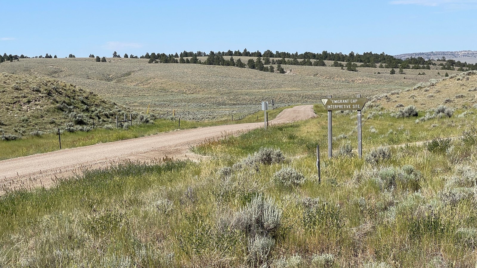 Desert with road between knolls, BLM sign, and distant wide sage hills beneath a pale sky.