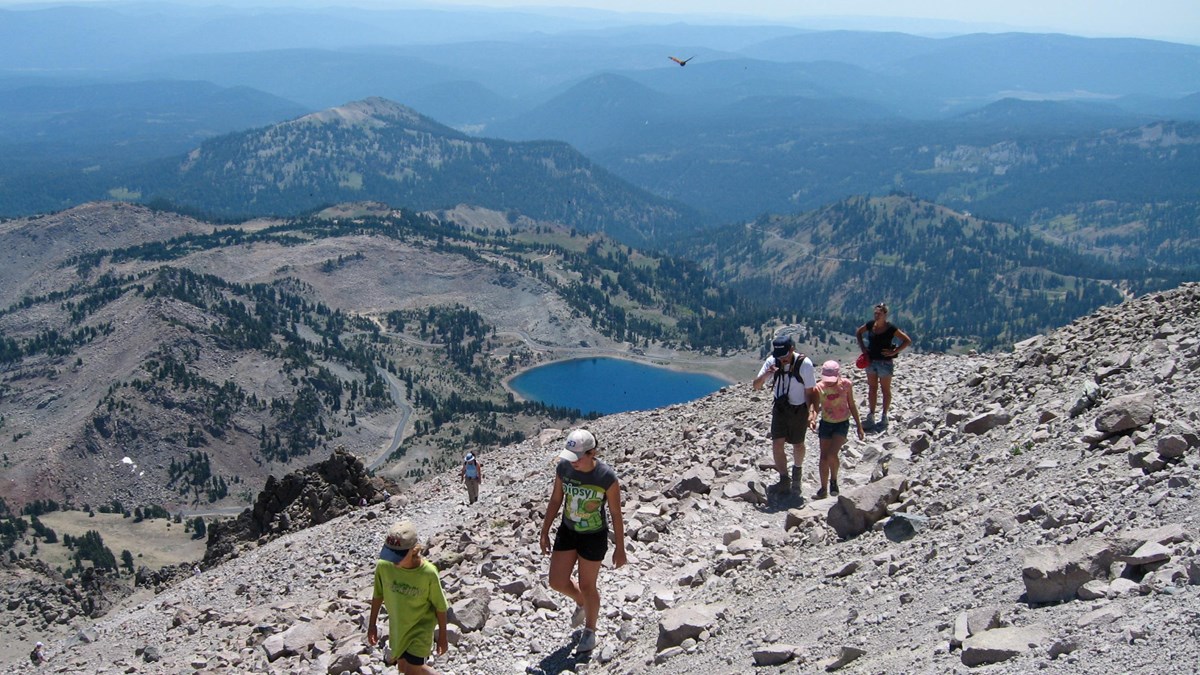 A family hikes up a rocky a trail above a bright blue alpine lake and distance volcanic peaks.