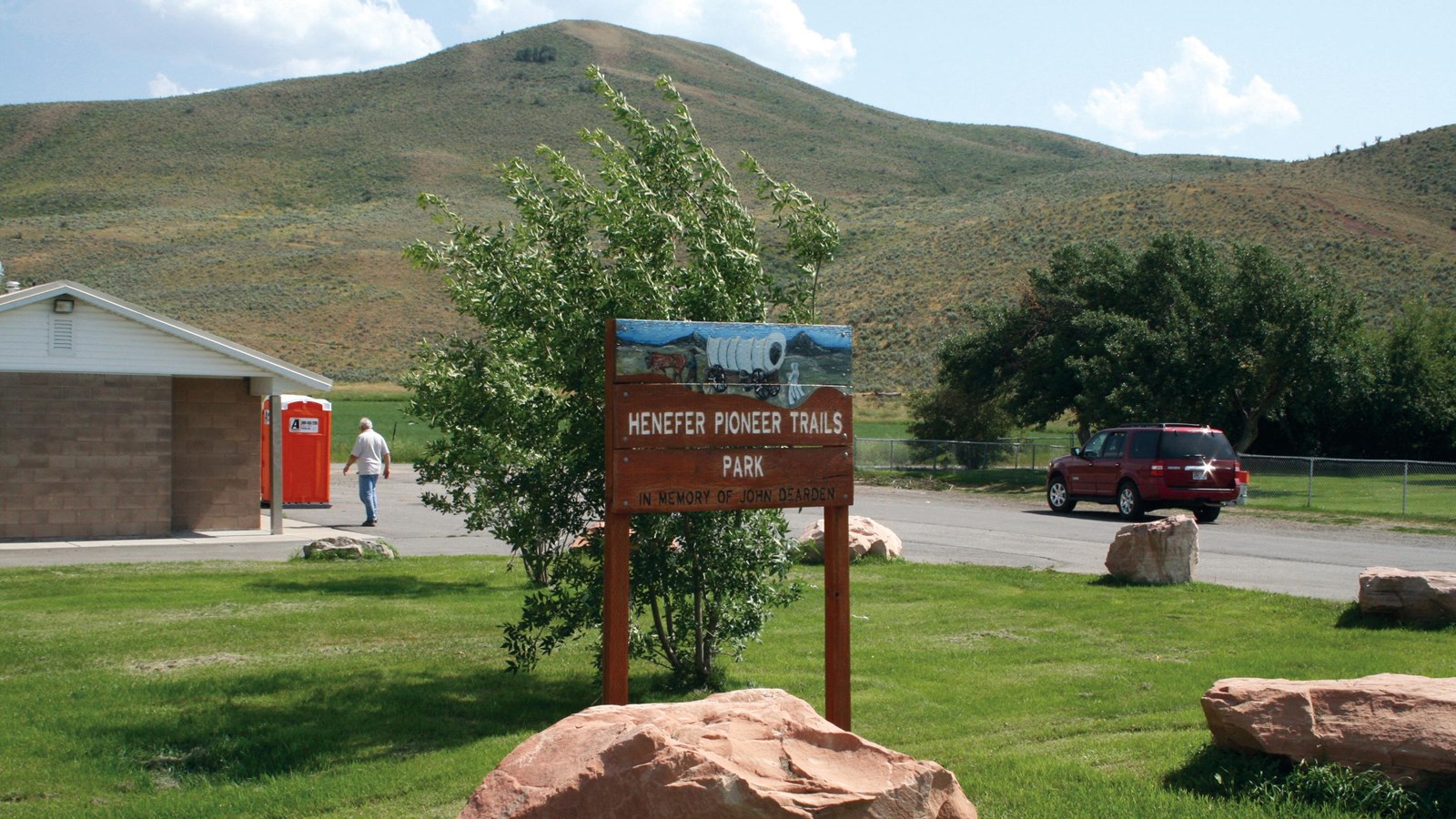 A brown sign sits in a grassy area in front of a small stone building and distant grassy hill.