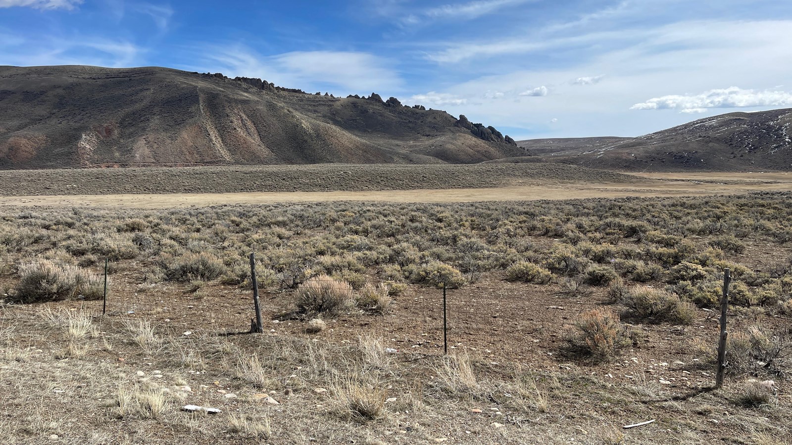 A fence, sagebrush plain, meadow, and steep hill lead to the jagged rock spine of The Needles site.