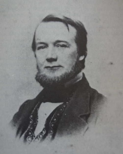 Portrait of a man wearing a dark suit with printed vest underneath. He has a short beard.