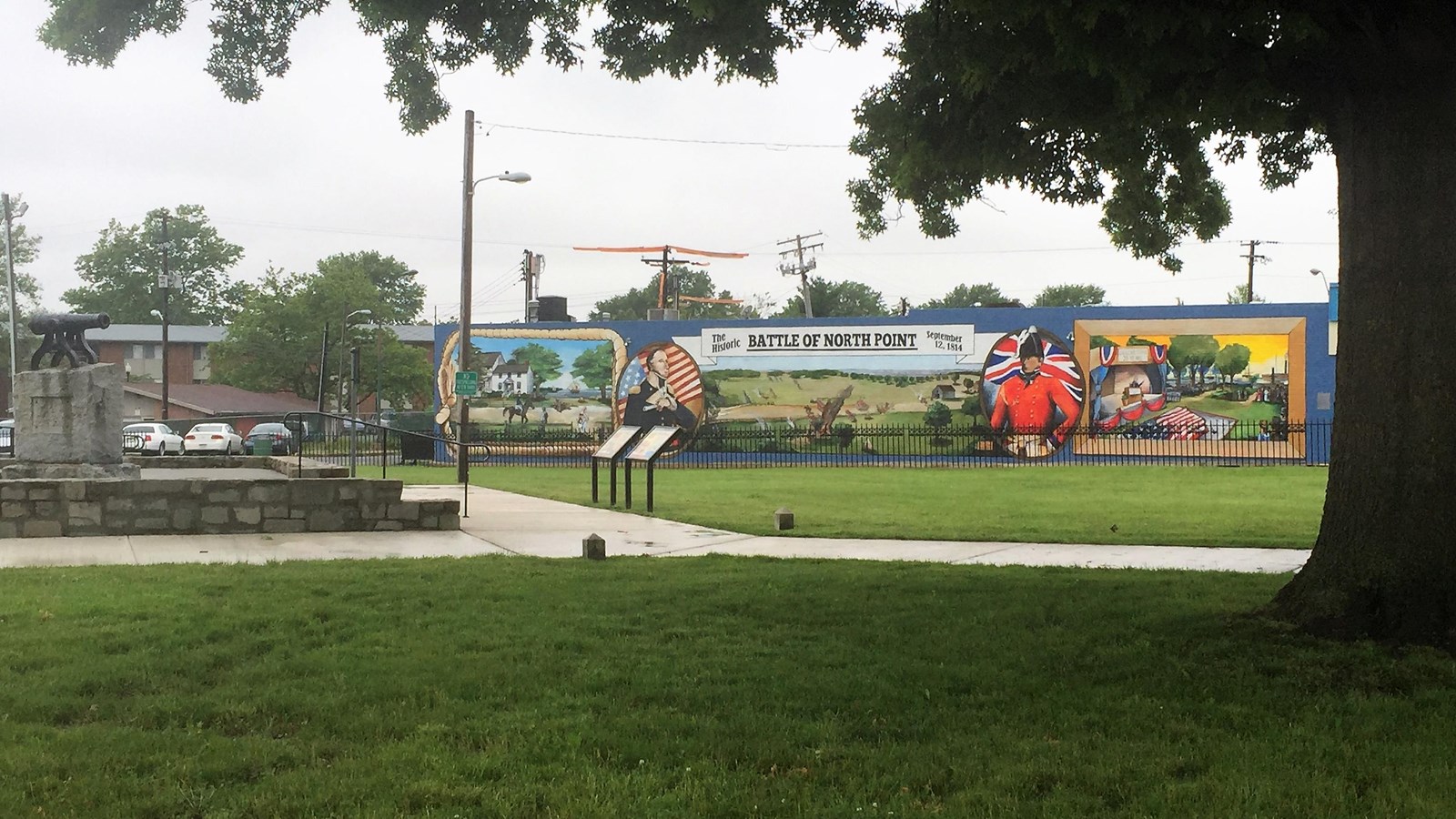 Small park with a tree in the foreground, cannon on the left, and a mural in the background.