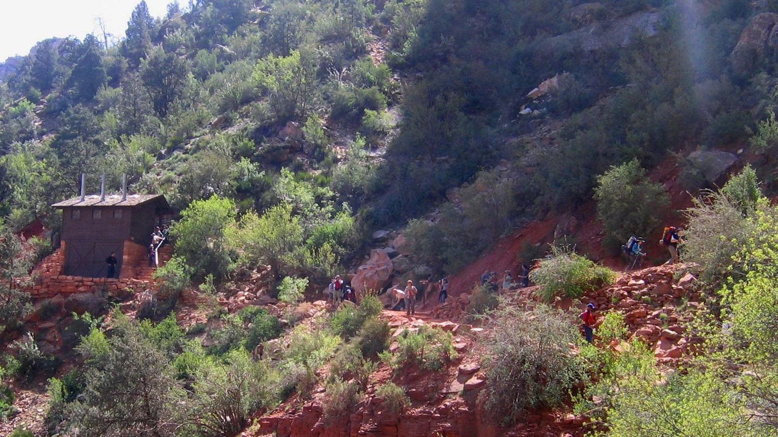 A small brown building on a steep slope of reddish rock covered by small bushes and trees.
