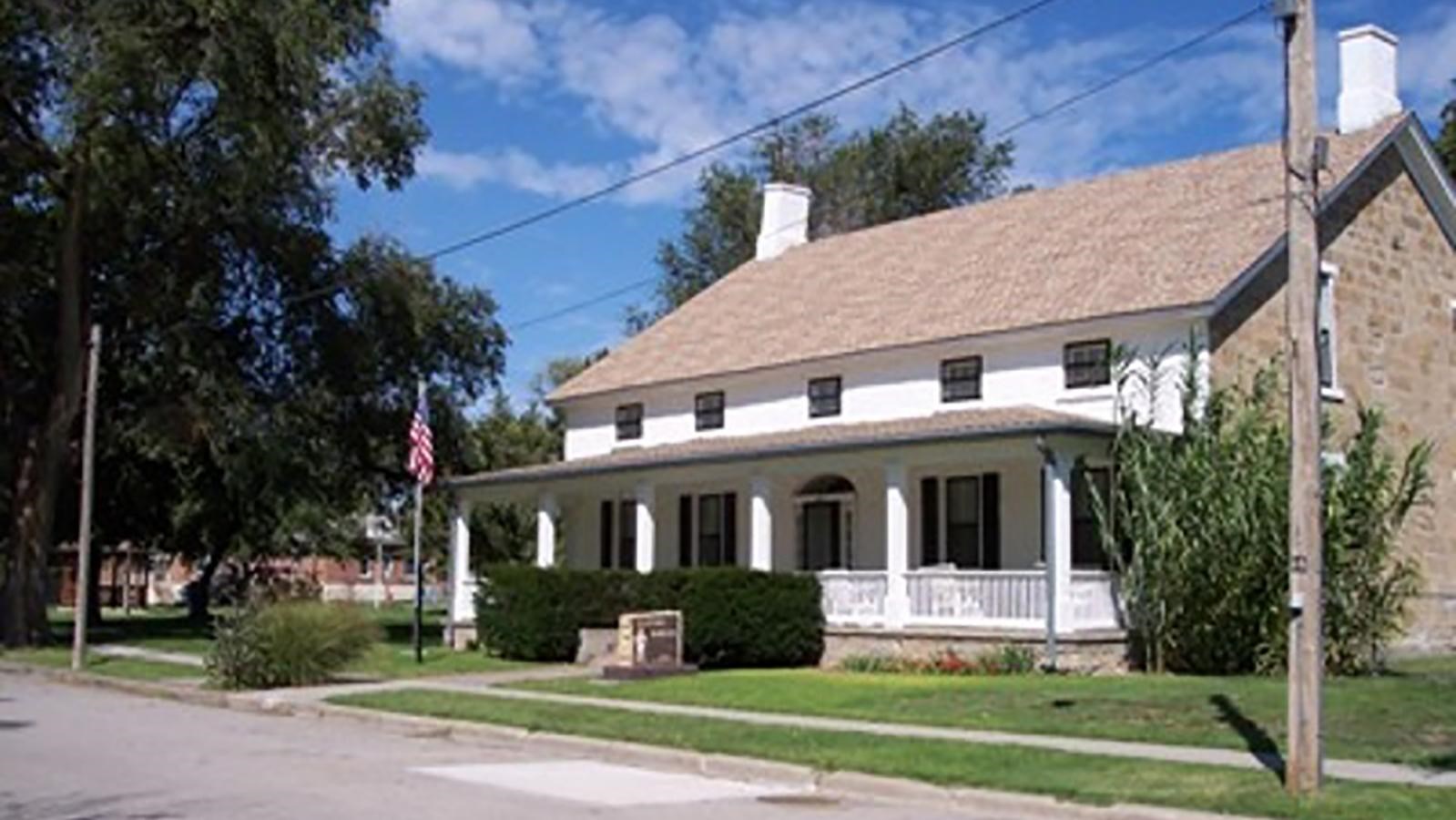 Stone building with porch and grassy lawn. Blue skies, trees, American flag also present.