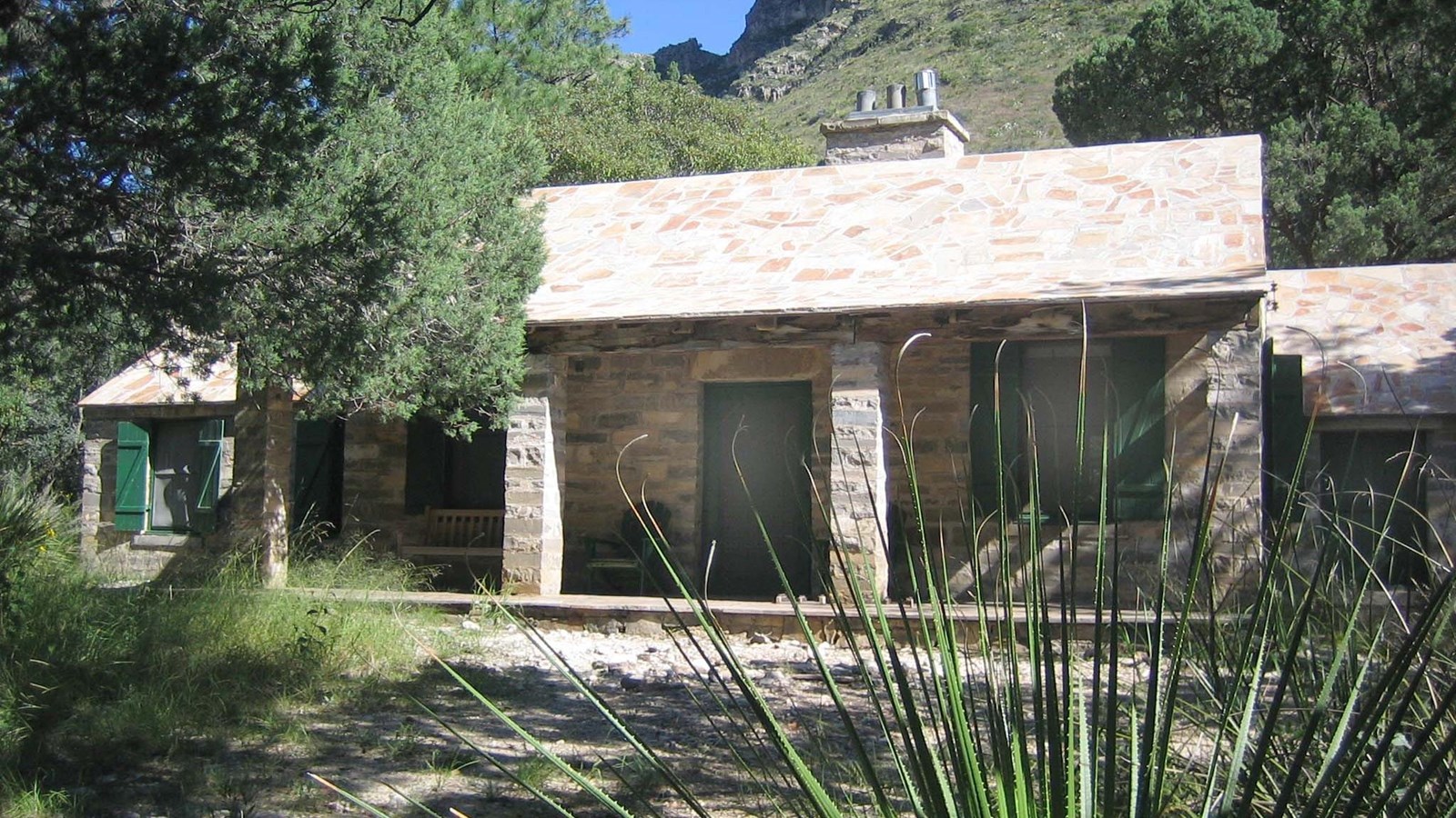 A cabin with stone walls and roof surrounded by desert mountain landscape and plants