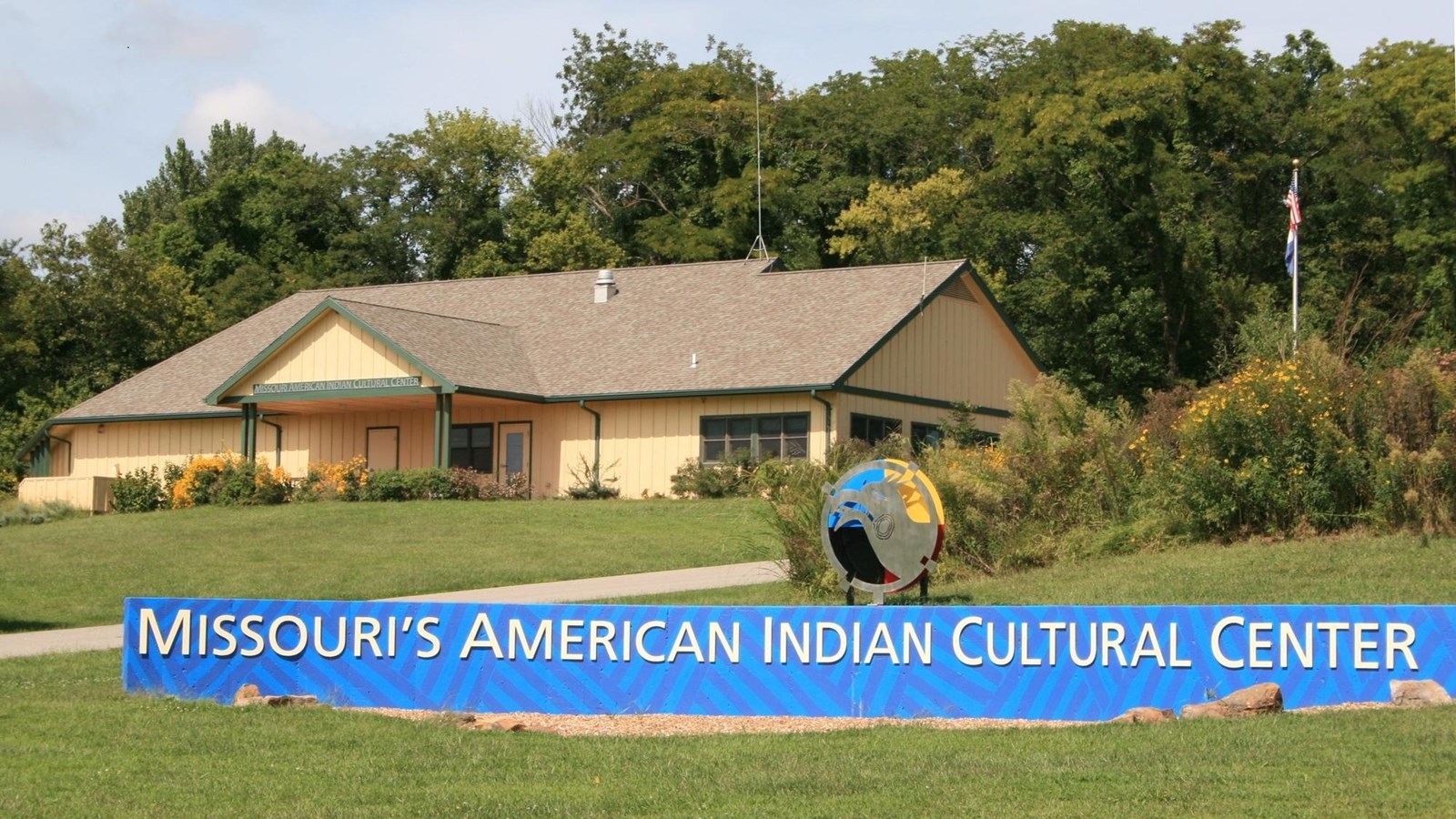 Blue sign reading, “Missouri’s American Indian Cultural Center” on a lawn in front a tan building