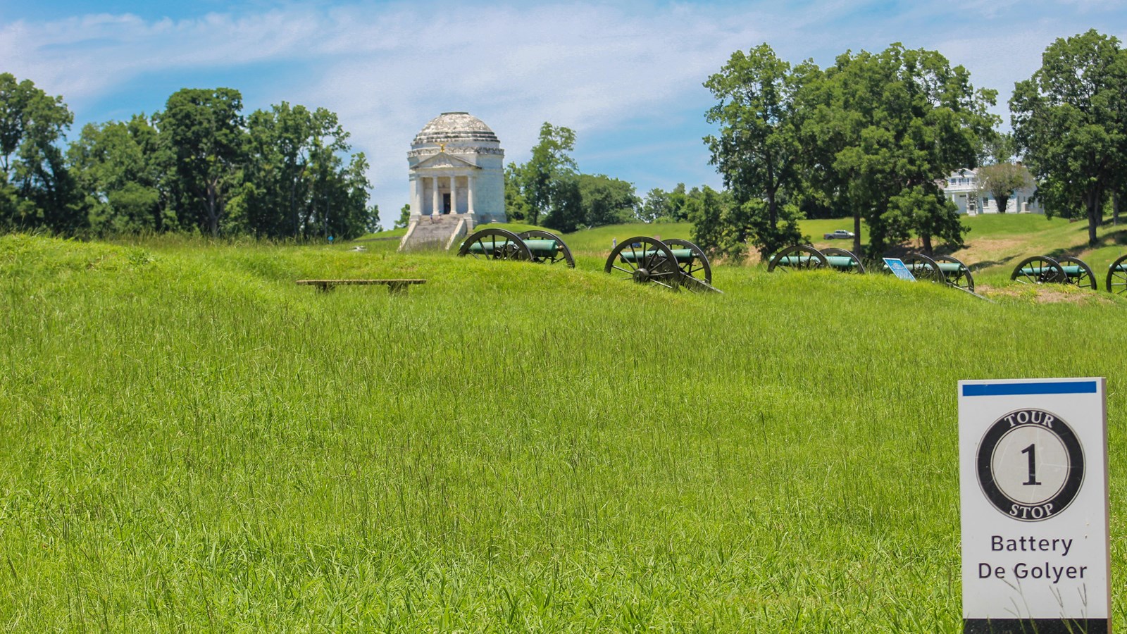 A tour stop sign next to a field with several cannons. In the distance is a large white monument.