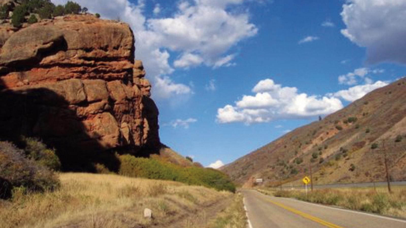 A large, red-hued rock outcropping hangs near a road through a canyon.