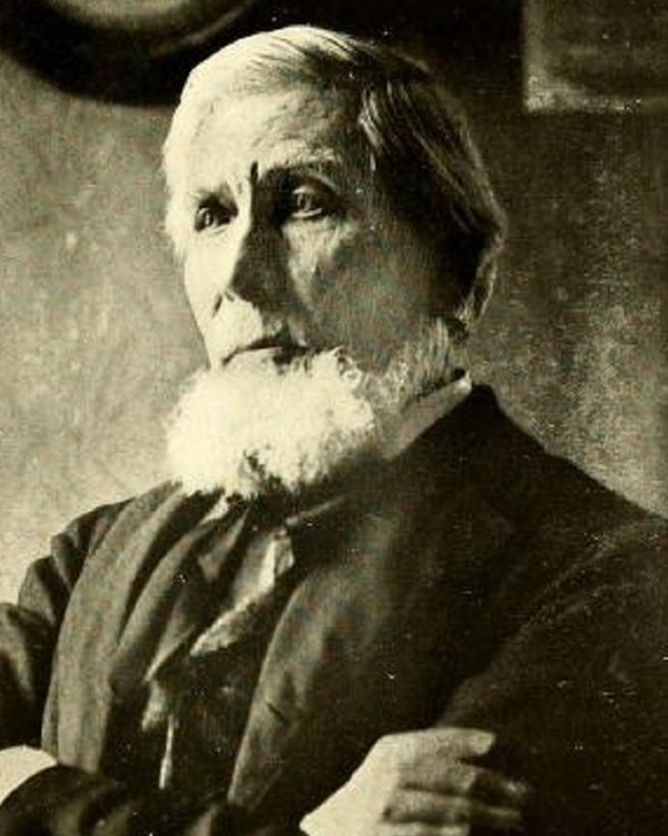 Portrait of a man with a long white beard and dark suit with his arms crossed over his chest. 