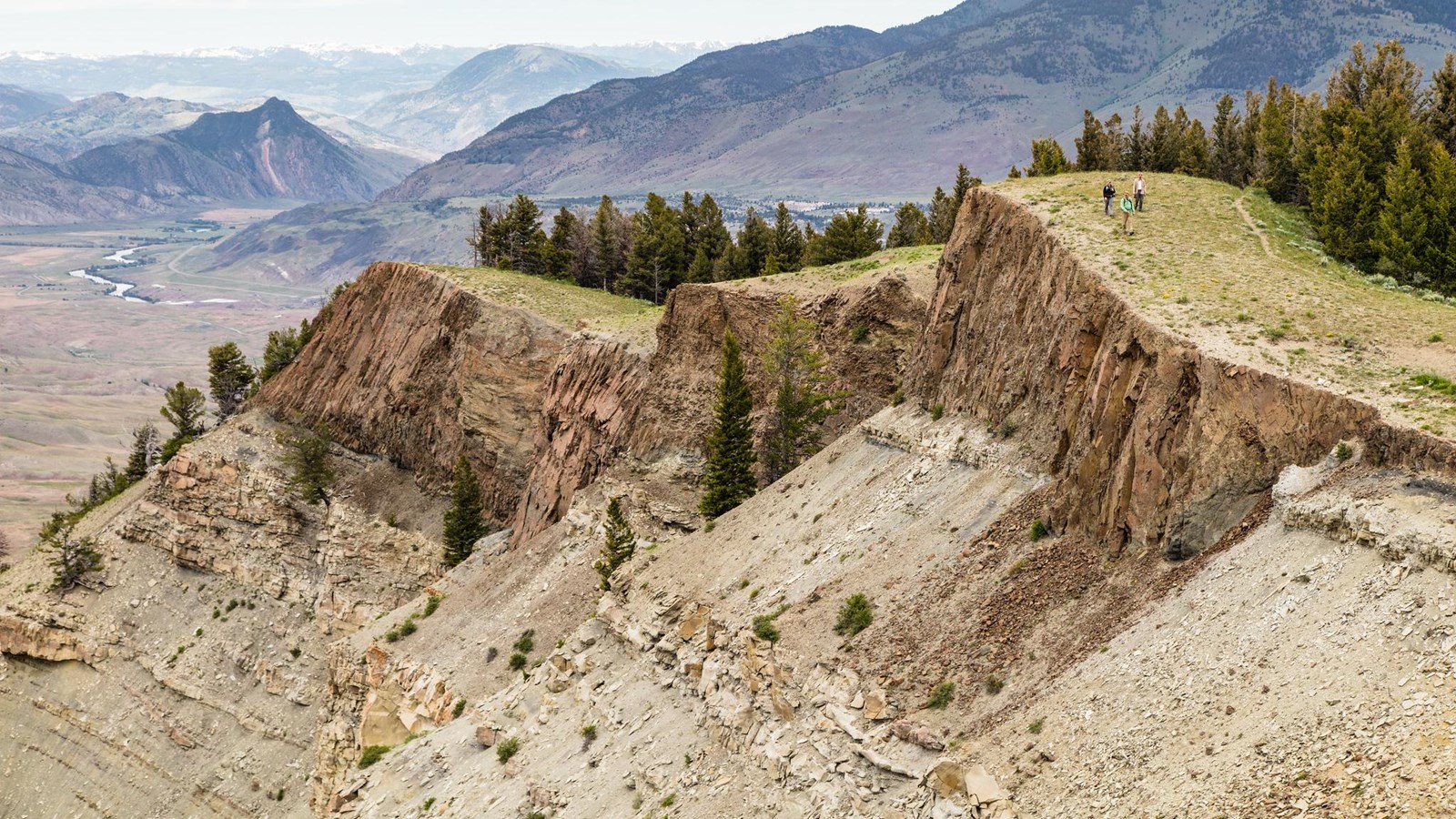 Three hikers walk on top of a mountain, with a river and grand mountains in the background.