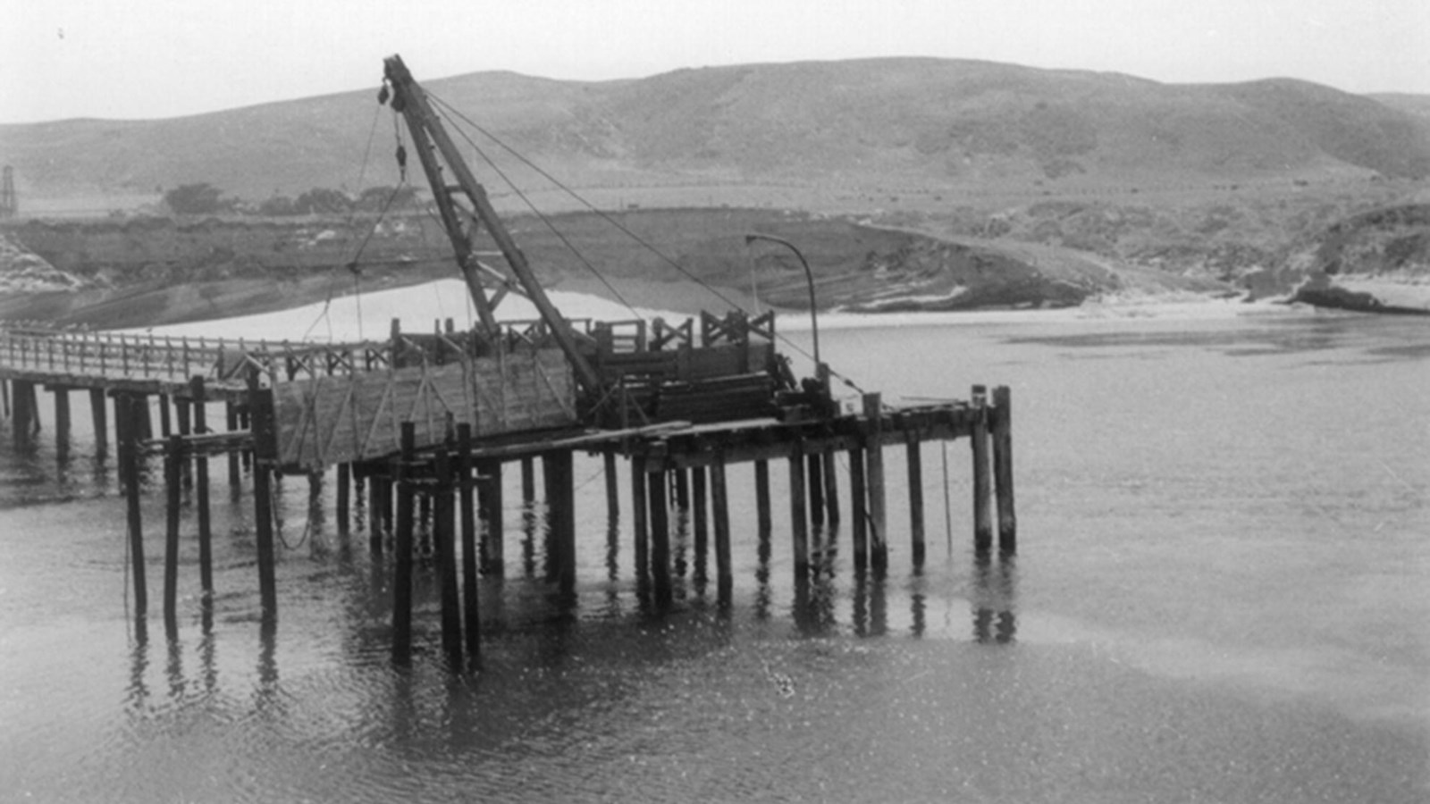 Large wood pier with wood cattle chute on end. Low hills and beach in background. 