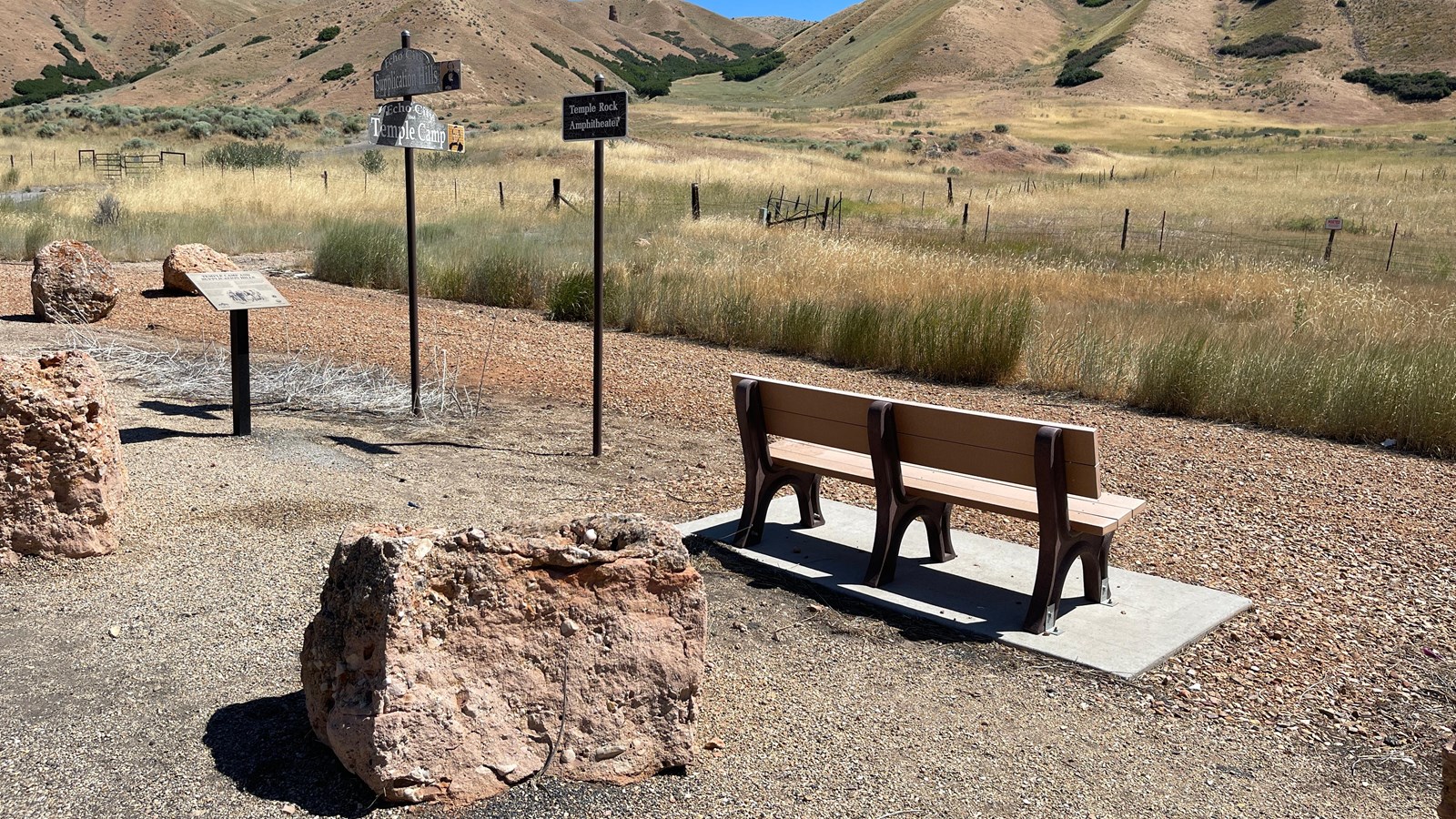 Boulders and a bench with signs near sit at foreground. Blue sky, mountains, and grass are above.