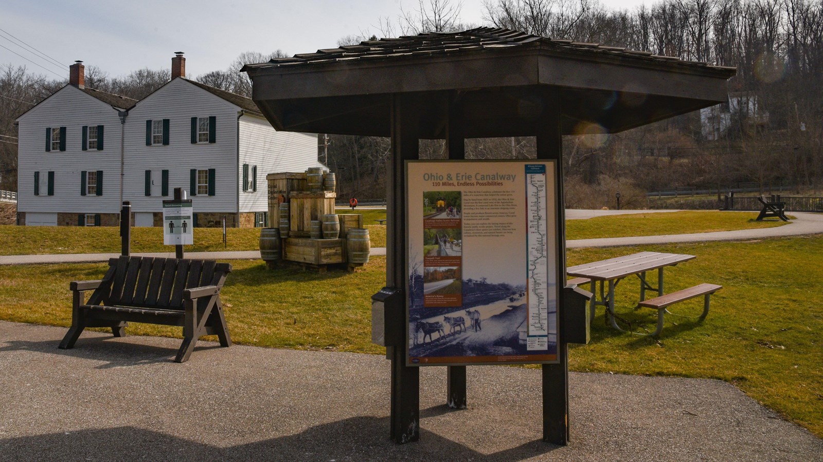 3-sided kiosk by a picnic table and benches. Path leads to a two-story white building behind.