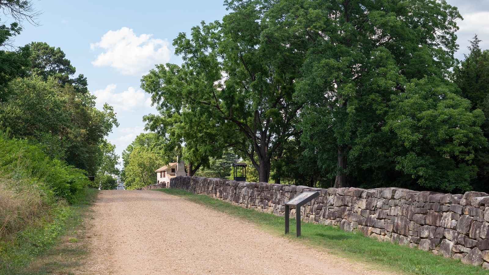 A gravel road lined by a stone wall with a small, two-story white house in the background
