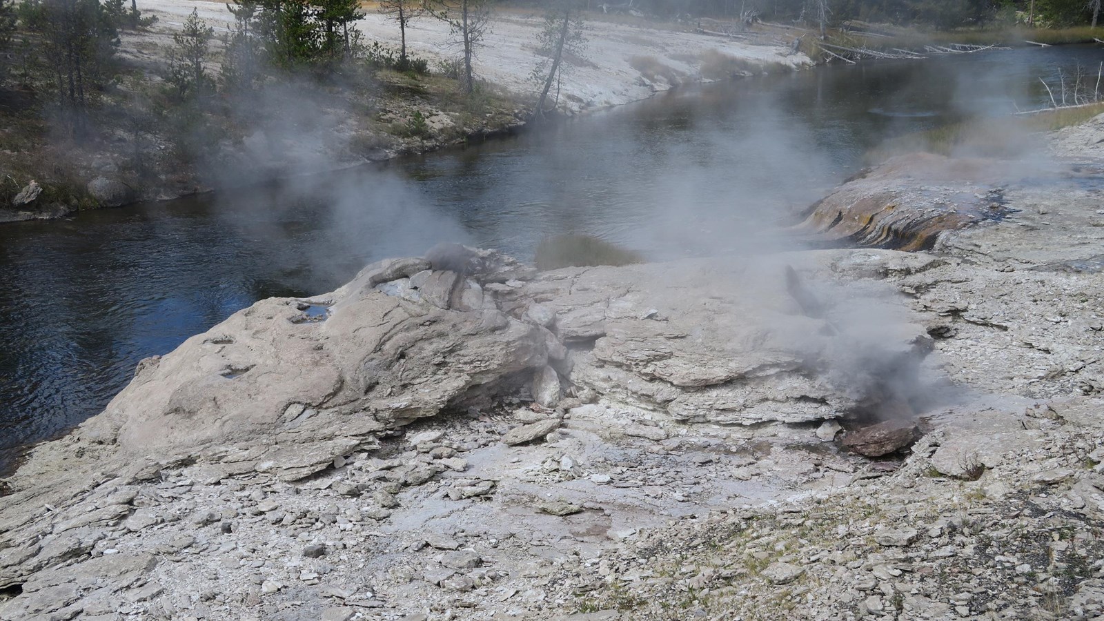 Steam rises from vents in layers of sinter on the edge of a river.