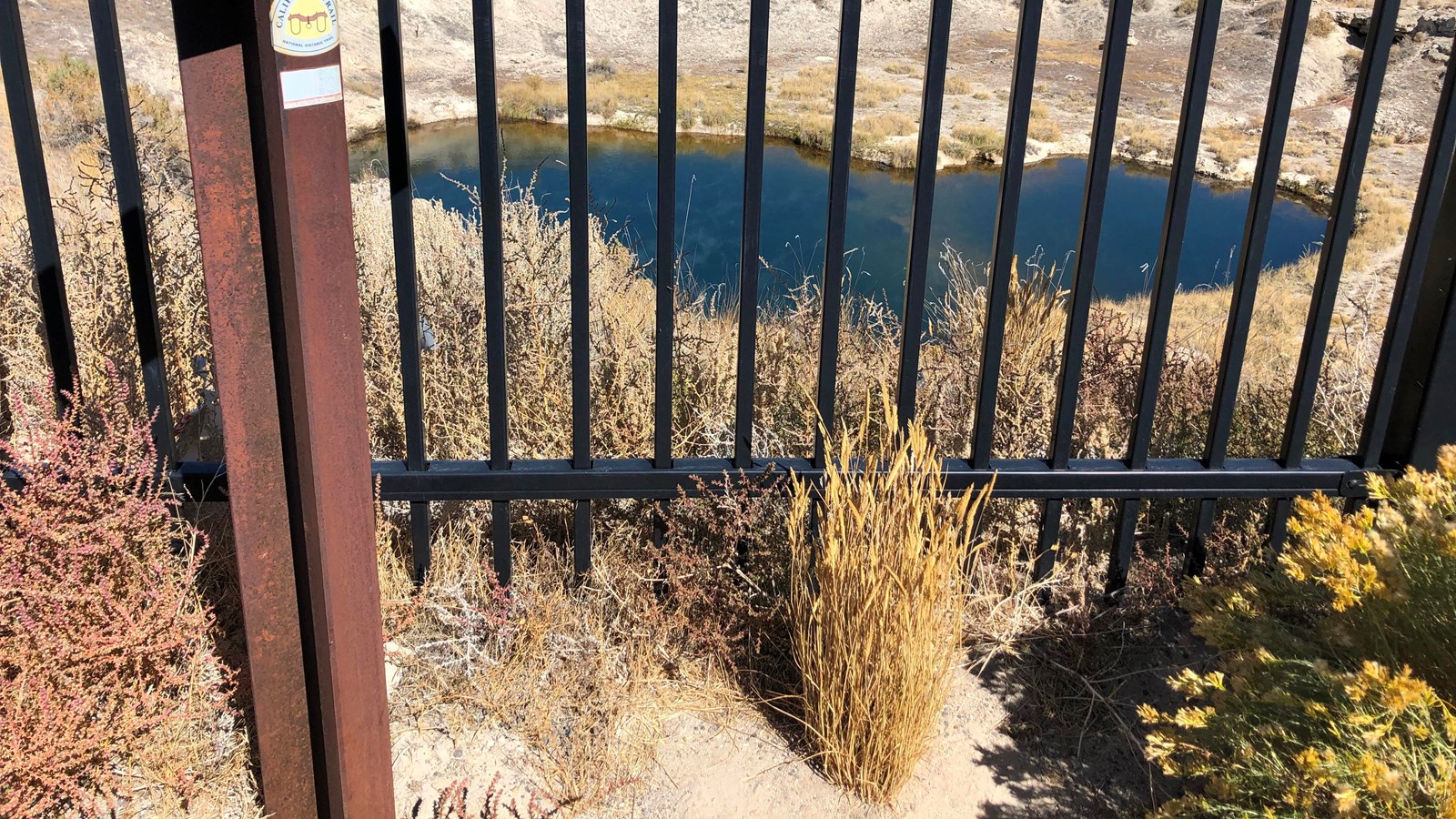 Steel bars block access to a small, deep blue desert pond.