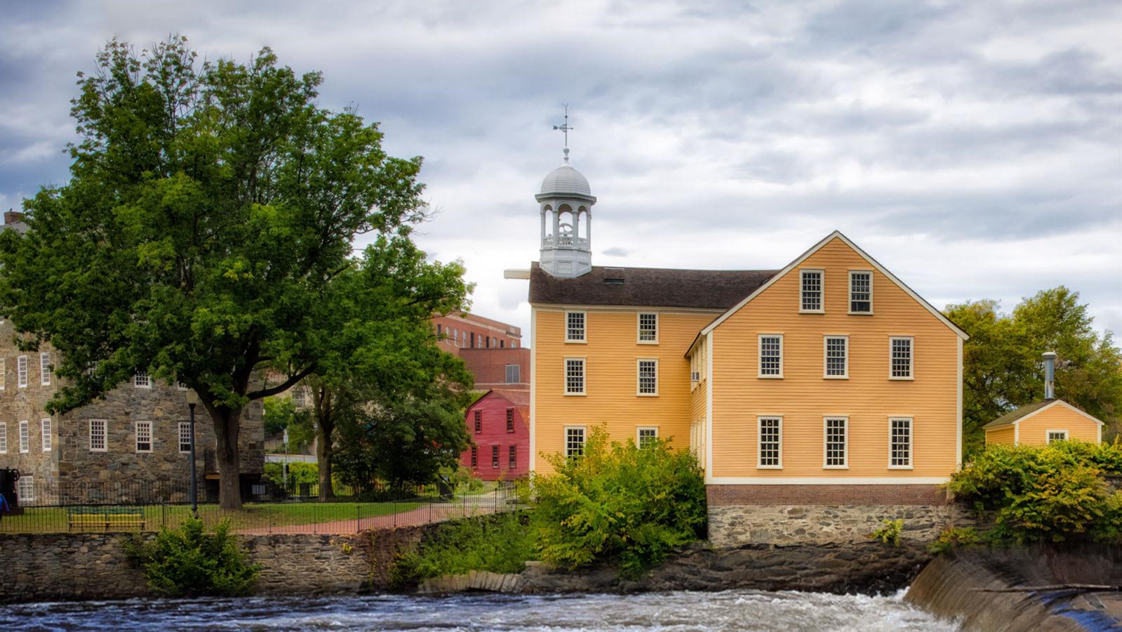 View across blue water to large yellow building and large stone building peaking out behind a tree.