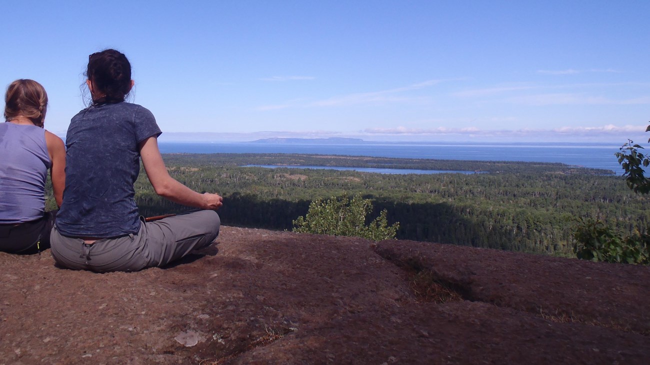 Two people sit on a rocky outcropping over looking a forest and lakes on an island in Lake Superior.