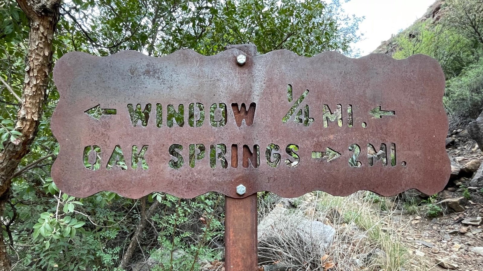 A brown metal sign has trail names and distances carved into it.