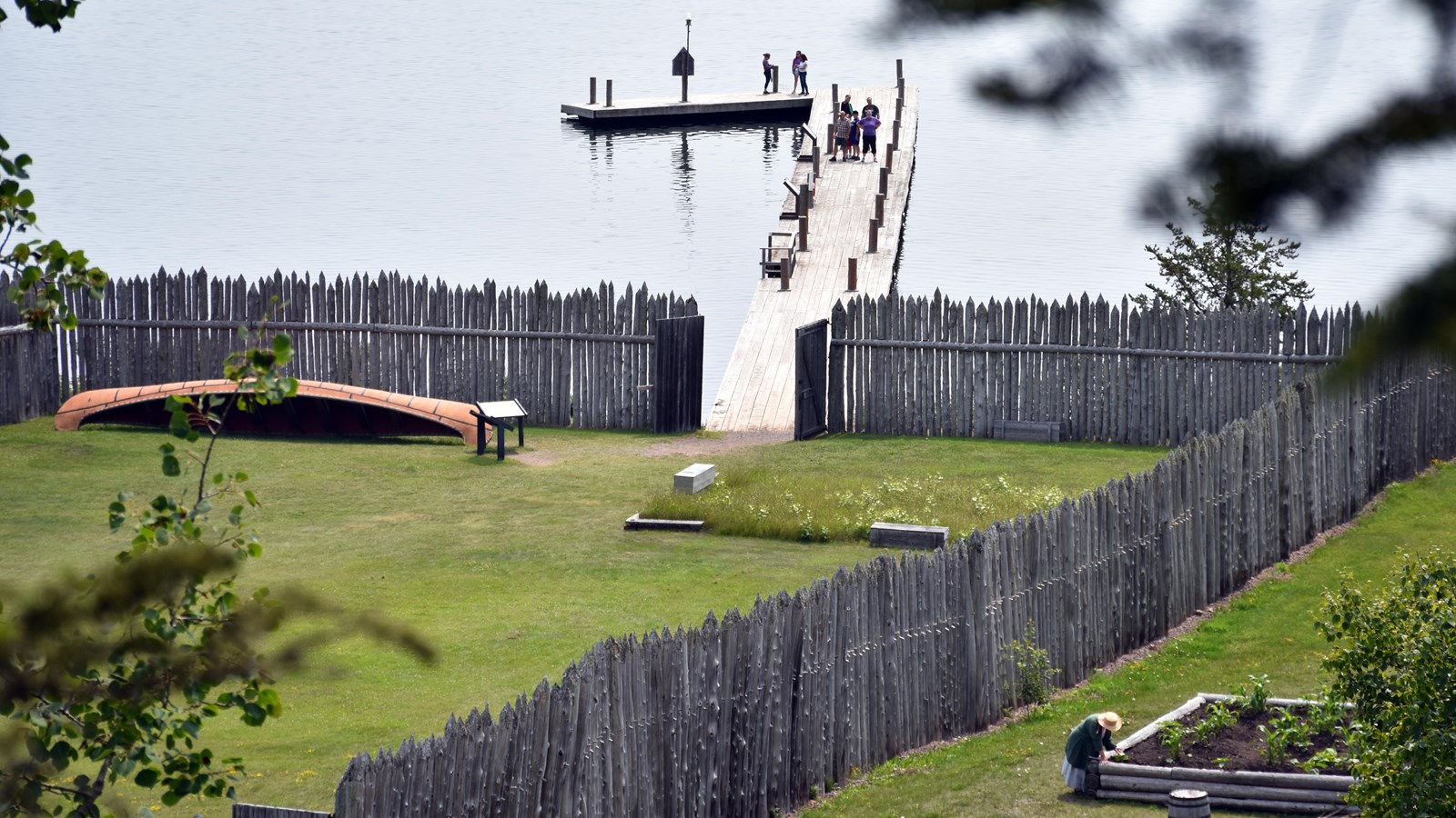 A person in historic clothing bending over a raised garden bed next to a stockade and nearby canoe.