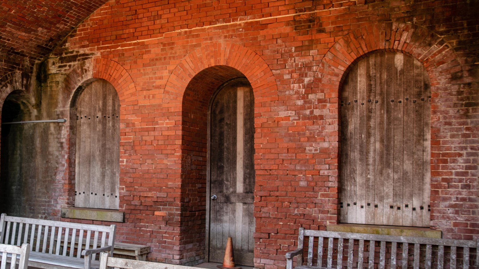 Wooden benches stand in front of a wooden door and two windows in a red brick fort.