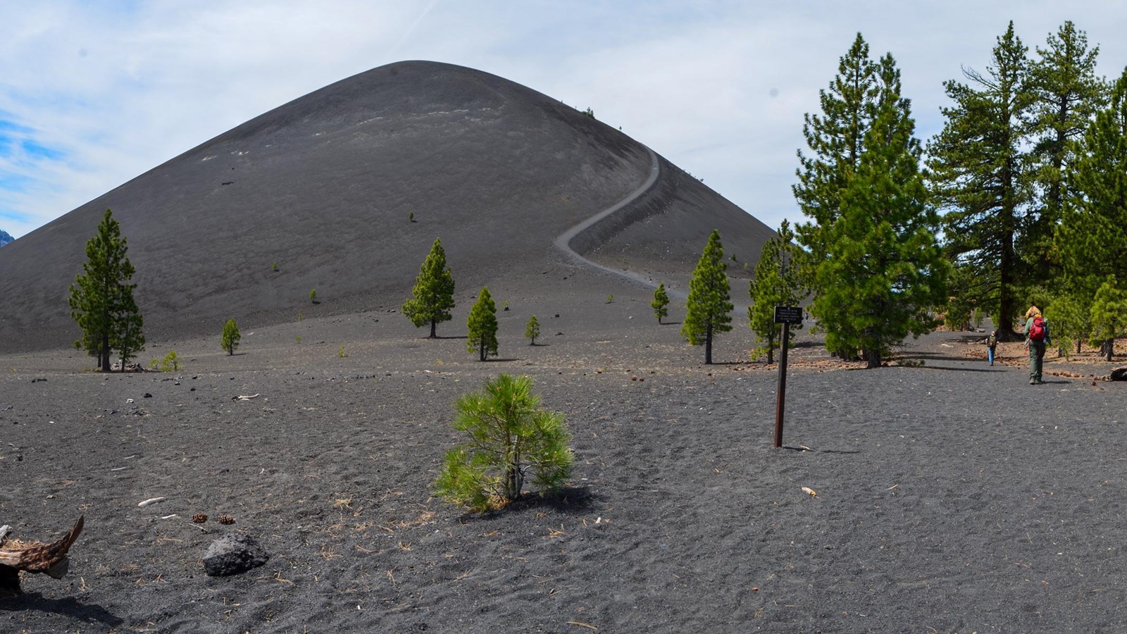 A panoramic view of a cinder cone volcano with sparse conifers at its edges. Two hikers walk on a su