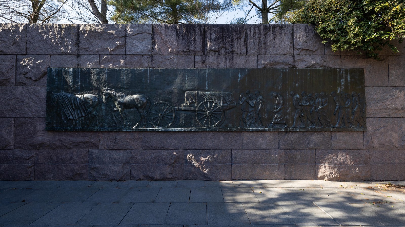 A bas relief is positioned on a stone wall. On the bas relief is a funeral cortege.