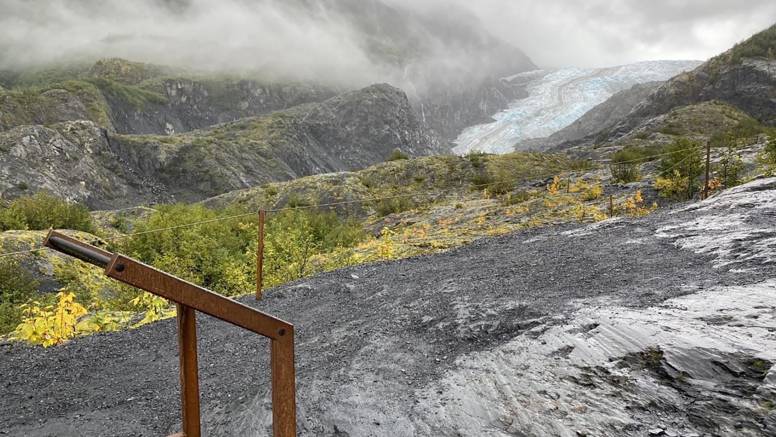 A sign in a metal case is in the front of the photo. The background is a glacier between mountains