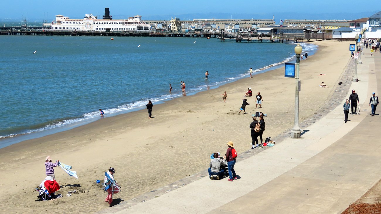 various people and children sit or play in the sand on the beach near Hyde Street Pier.