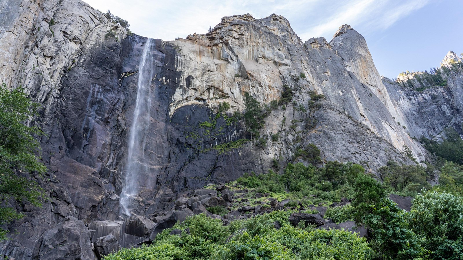 A tall, wispy waterfall cascades down a sheer rock face framed by moss and trees