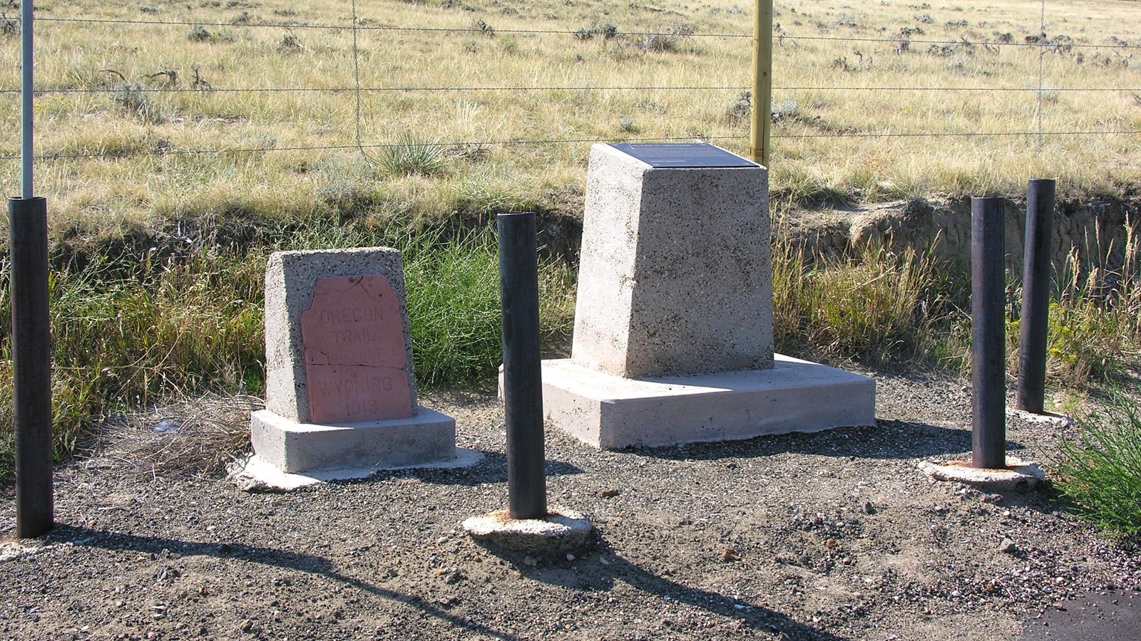 Two concrete roadside monuments with posts and plaque stand before a grassy ridge and fence.