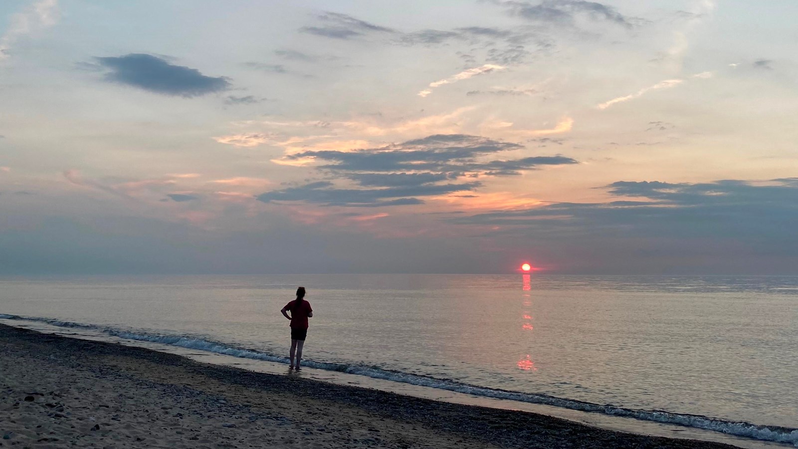 View of Lake Michigan beach at sunset with a hazy red sun on the horizon with a blue cloudy sky