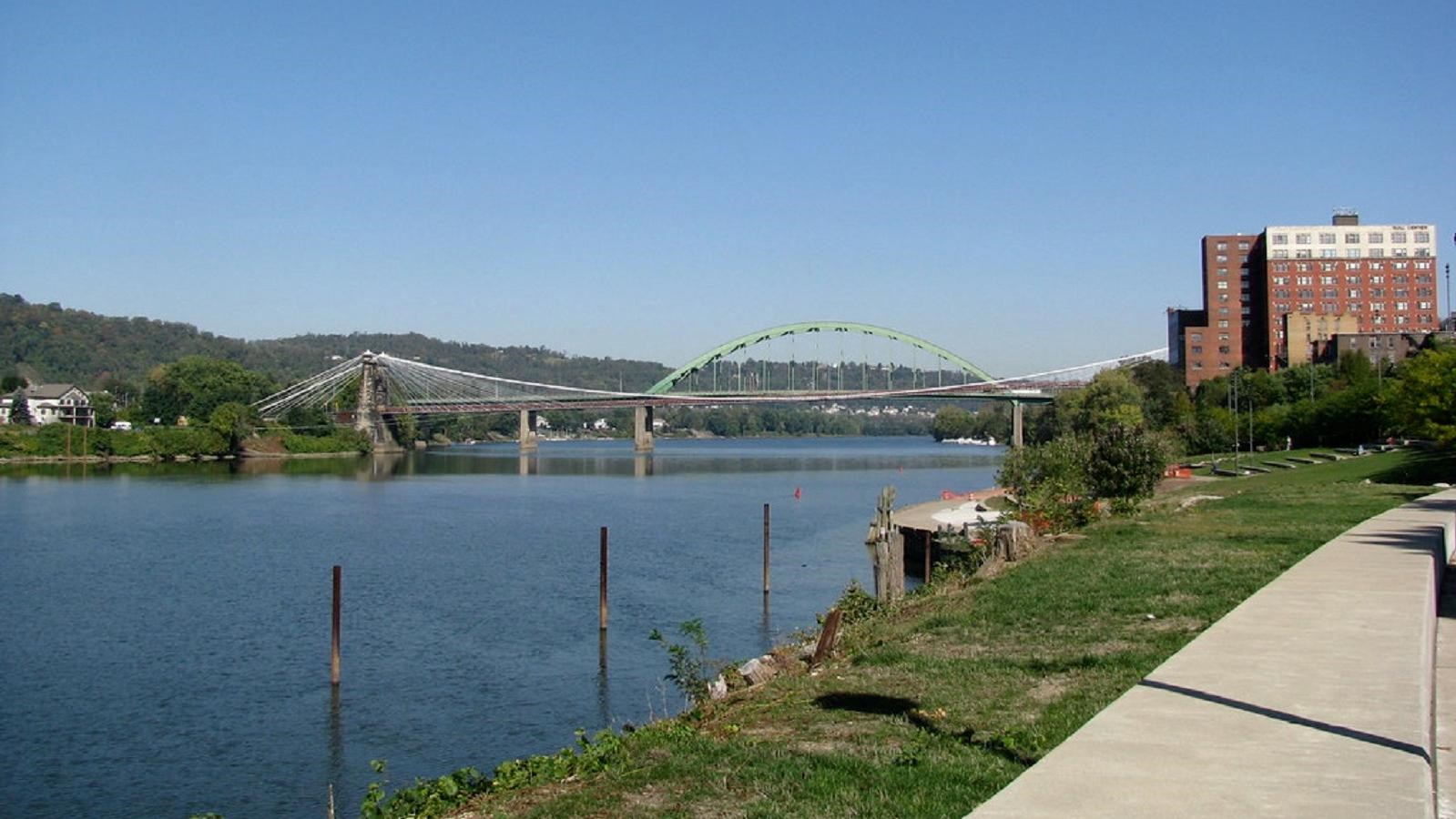 A suspenion bridge crosses a wide river, stretching from one grassy bank to the other