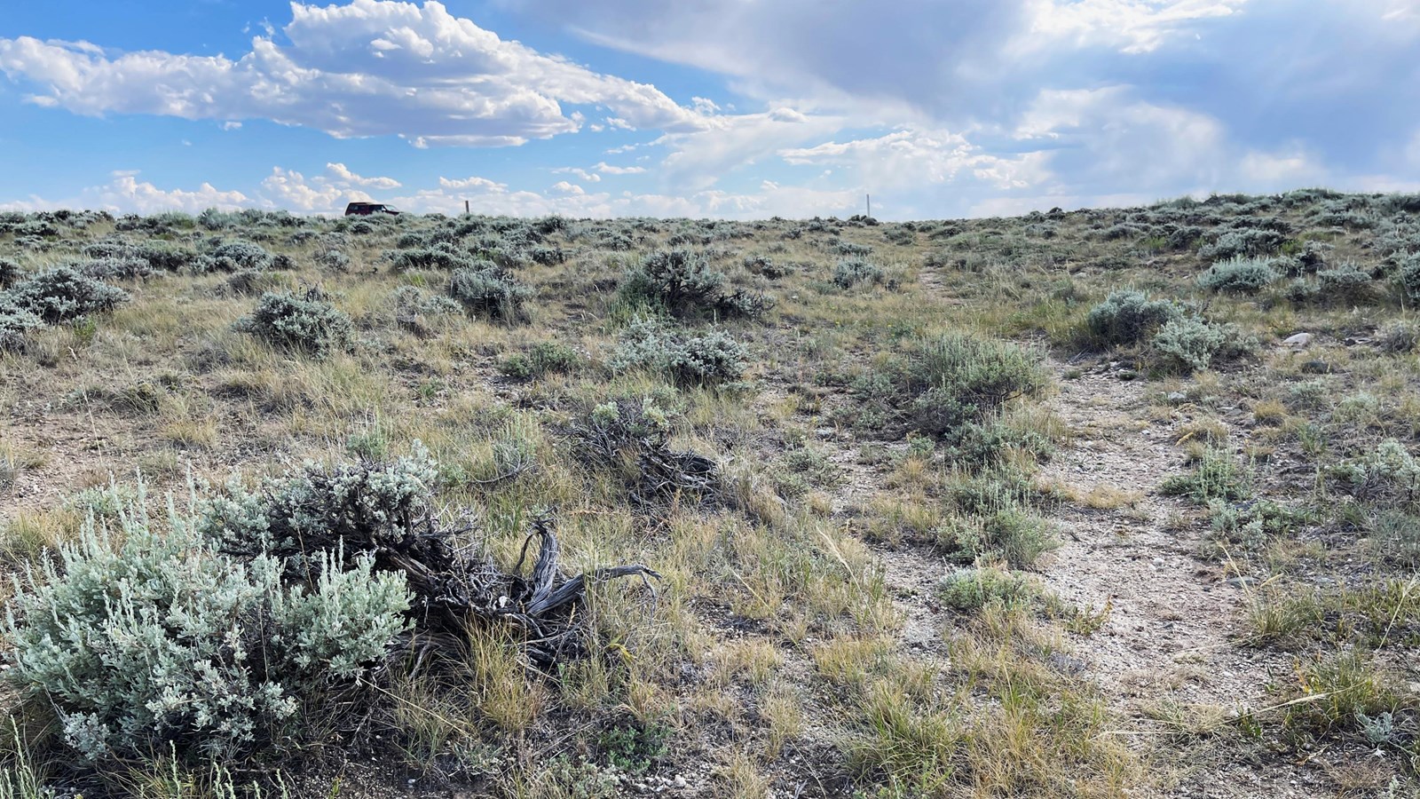 A gravel path climbs a sagebrush hill with trail markers and a truck below underneath a cloudy sky.