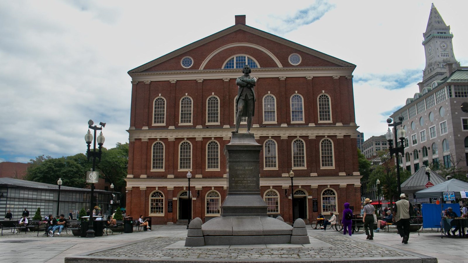 A statue stands amongst people in the middle of a square in front of a large brick hall.