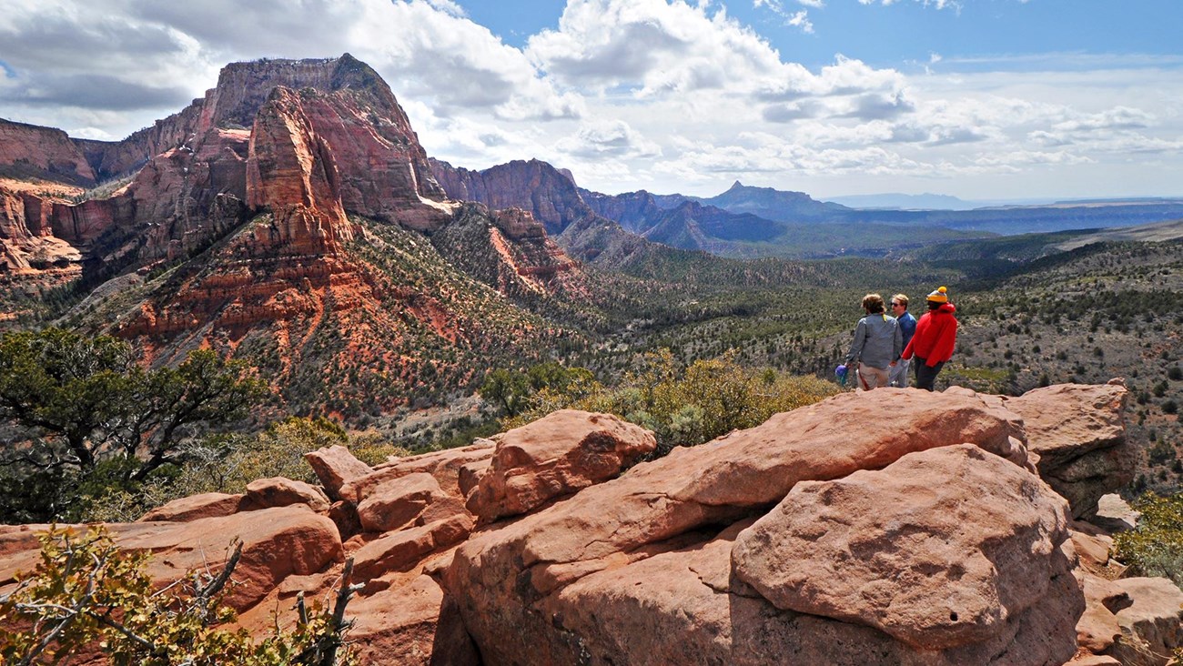 Red sandstone rocks with three hikers standing on them looking out over a valley. Blue, cloudy sky.