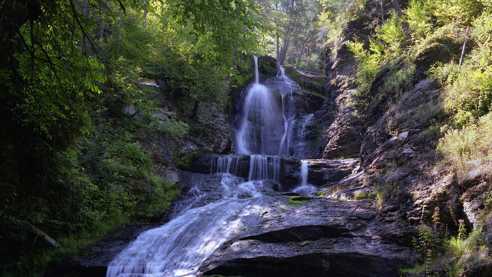 A waterfall in a ravine with rock ledges and green foliage surrounding it.