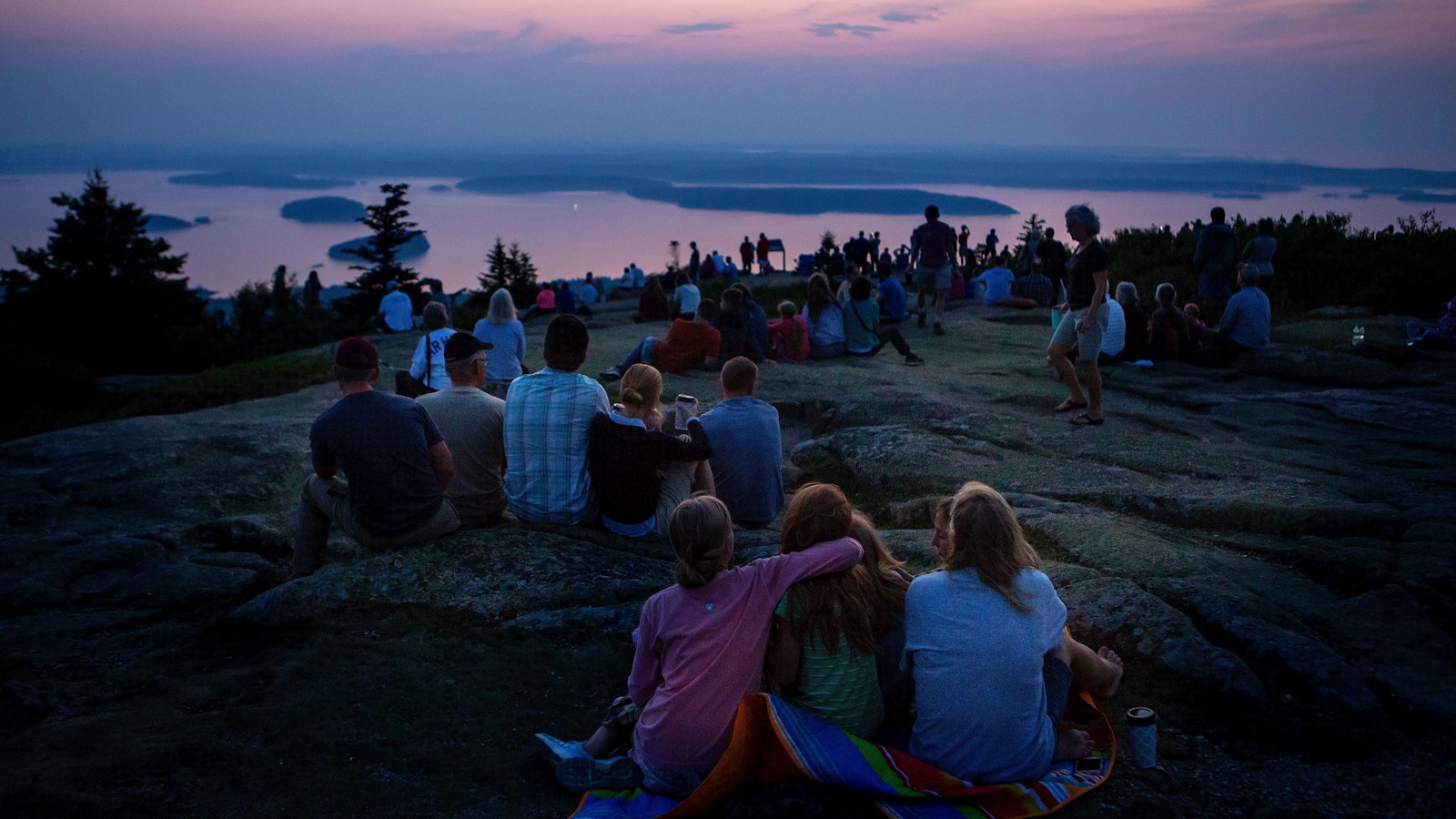 Groups of people sit on a granite outcropping, looking out at a view of sunrise over ocean.