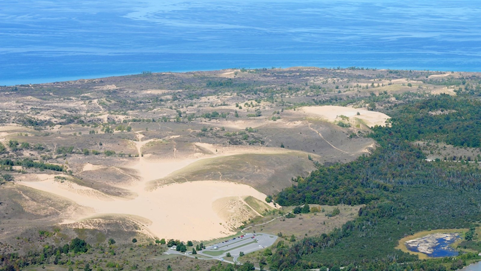 Aerial view of sand dunes and surrounding forest. Lake Michigan is visible in the background.