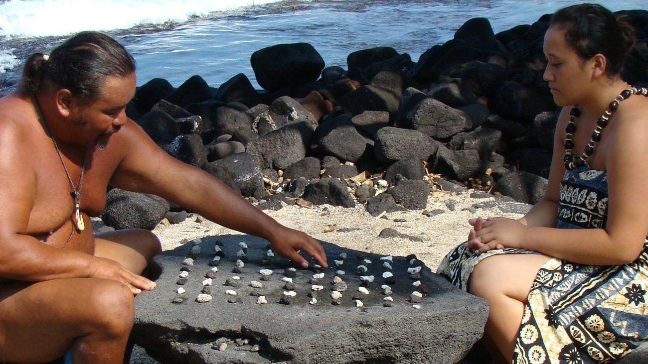 A man and and woman in traditional clothing sit at a kōnane board. The man reaches for a game piece.