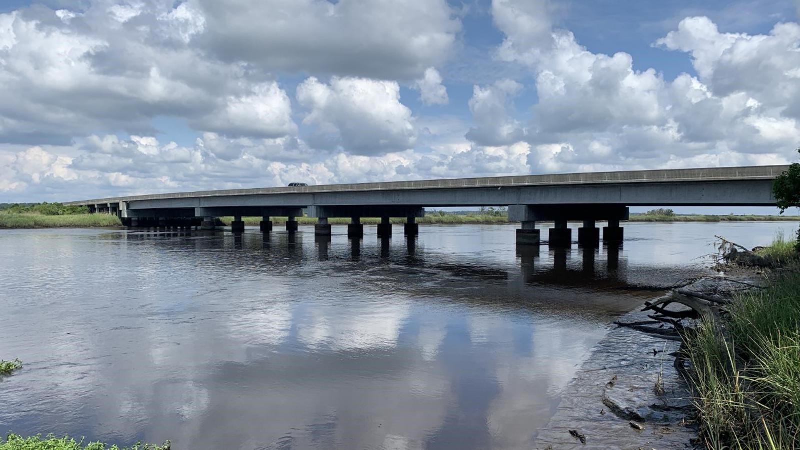 A concrete bridge spans over a wide river with grass on the shore and clouds in the sky.