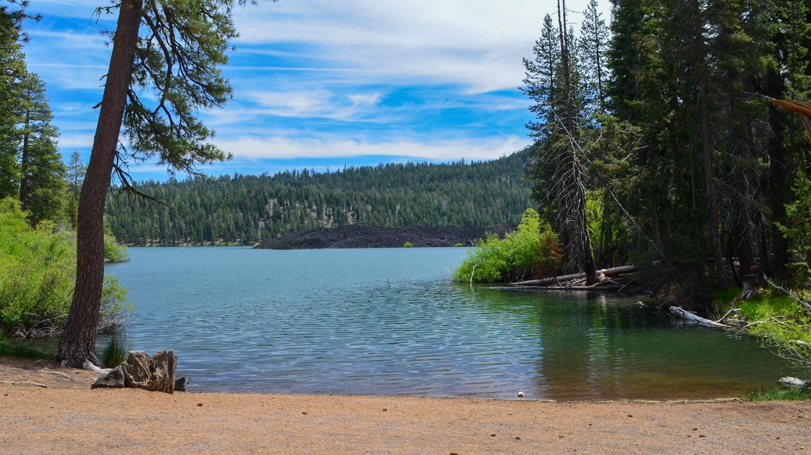 A gravel boat launch area at the edge of a large lake lined by conifers and bushes.